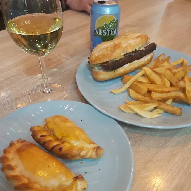 Una comida compuesta por sándwich, papas fritas y empanadas, acompañadas de una copa de vino blanco y una lata de Nestea, en una mesa de madera clara.