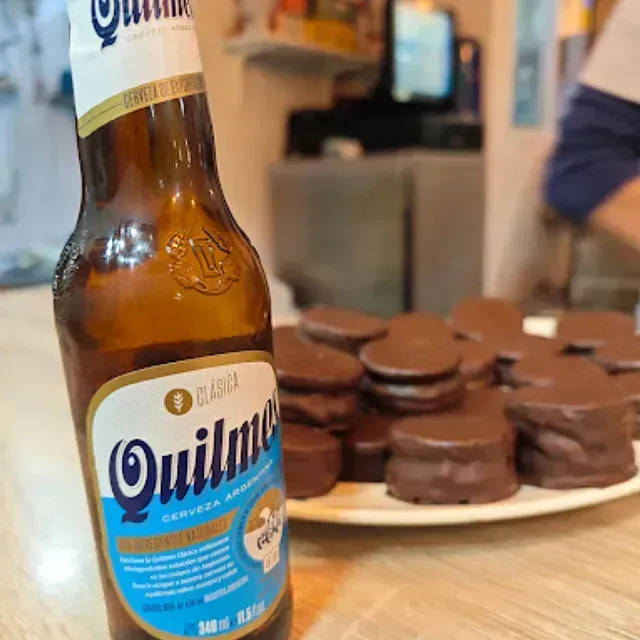 Una botella de cerveza Quilmes junto a un plato de alfajores cubiertos de chocolate sobre una mesa. Fondo borroso.