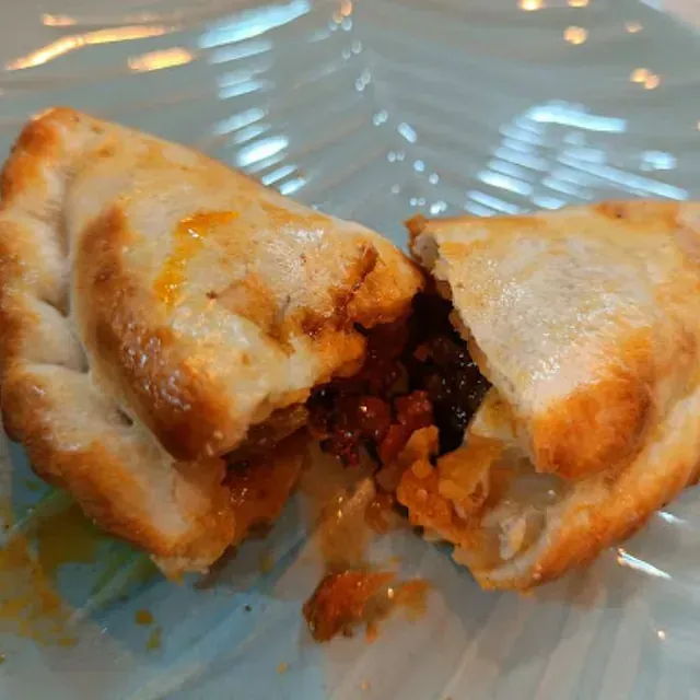 Una empanada cortada por la mitad, con un delicioso relleno, reposando sobre un plato azul claro. La corteza es dorada y el relleno se ve oscuro y jugoso.