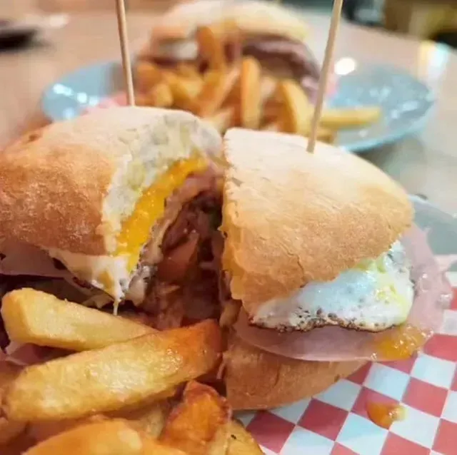 Un sándwich partido a la mitad con huevo, jamón y papas fritas en un plato a cuadros, otro sándwich y papas fritas en el fondo.