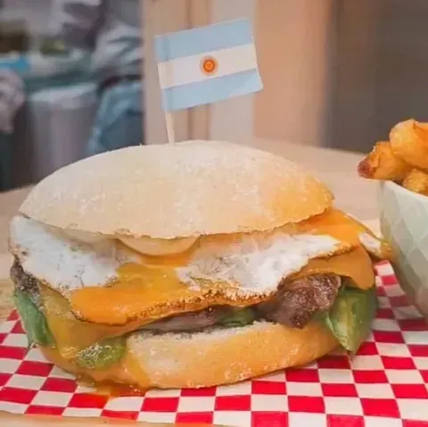 Hamburguesa con huevo frito, queso y palillo con la bandera argentina, servida en papel cuadriculado.