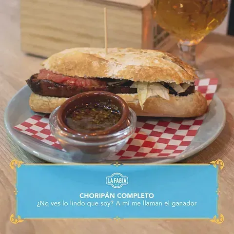 Sándwich de choripán con salsa para mojar en un plato, con un vaso de cerveza en el fondo y un texto azul con el logo de un restaurante.