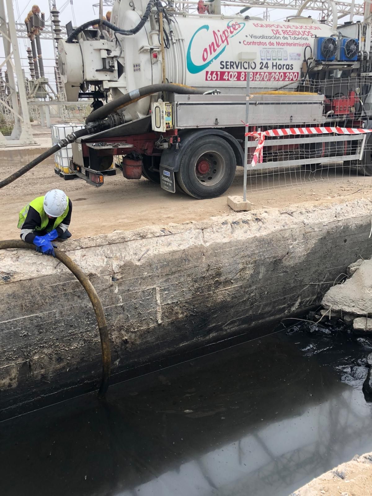 Un trabajador se agacha junto a un canal de drenaje, al lado de un camión de servicios públicos, e inspecciona una manguera cerca del agua.