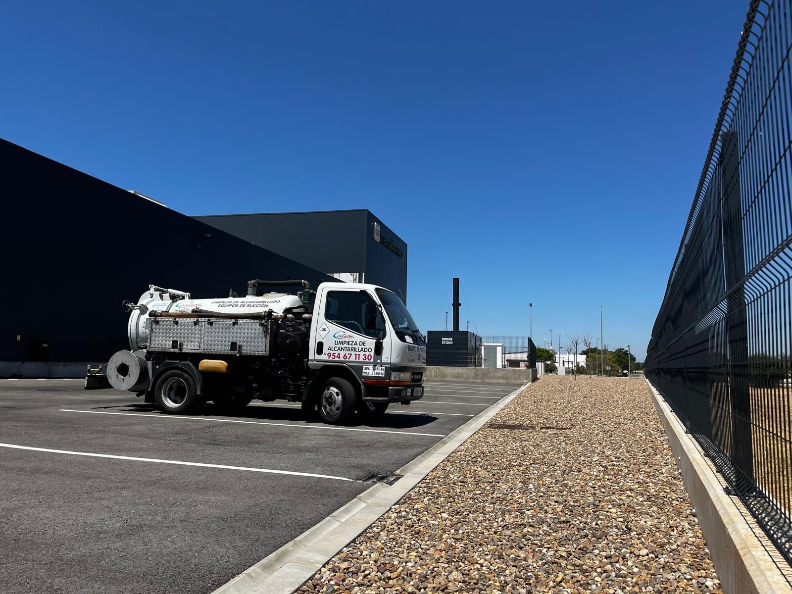 Camioneta blanca estacionada en un lote industrial junto a un camino de grava y una cerca de alambre bajo un cielo azul despejado.