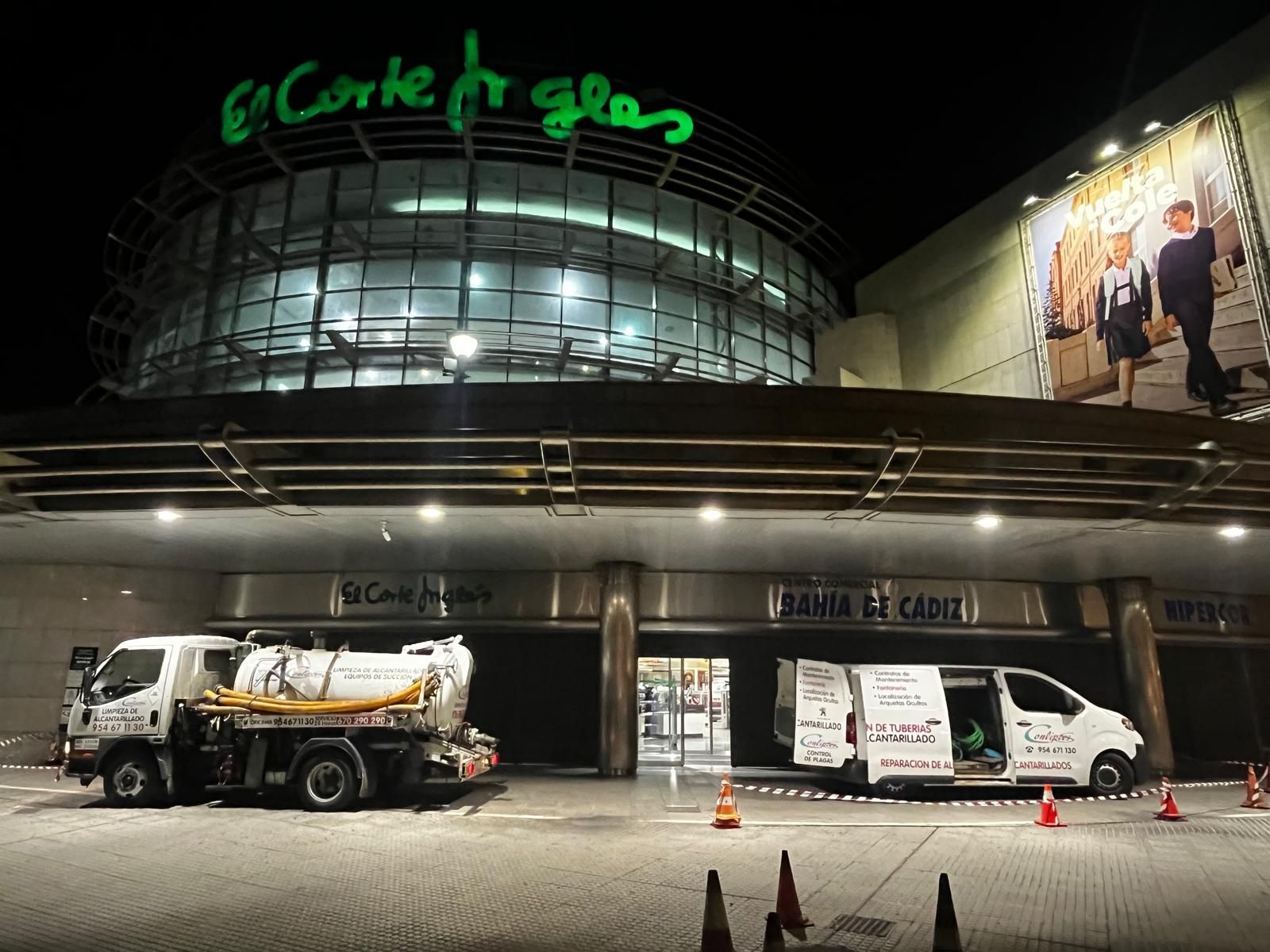 Vista nocturna de la fachada de una tienda con un letrero de neón verde, un camión de servicios públicos estacionado y una furgoneta bajo luces brillantes.