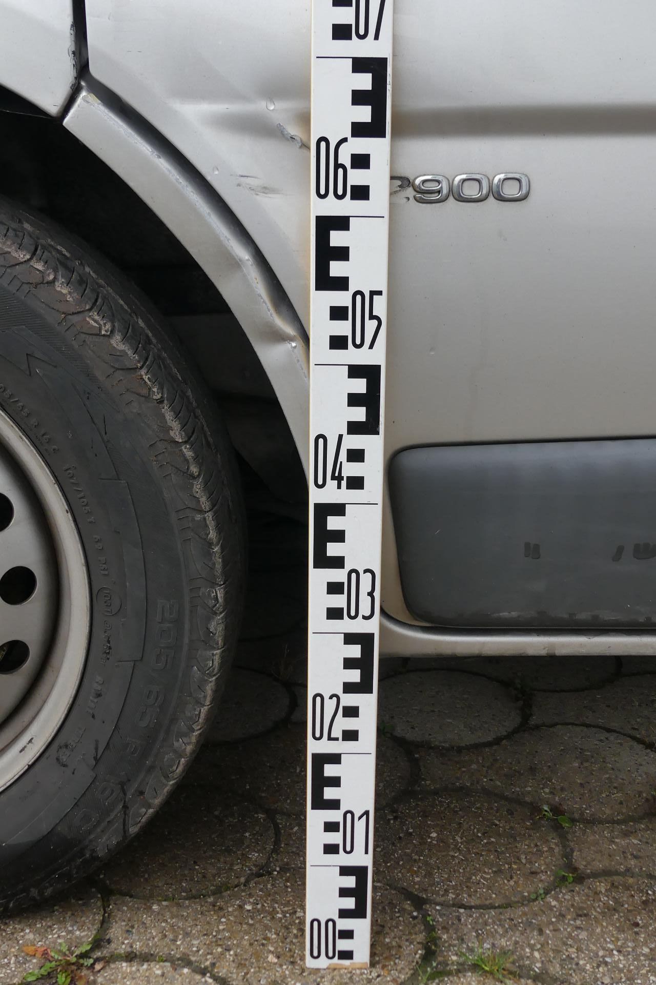 Nahaufnahme eines silbernen Autos mit einer beschädigten Seite, auf der neben einem Reifen ein Standardmaßstab für Messungen zu sehen ist.