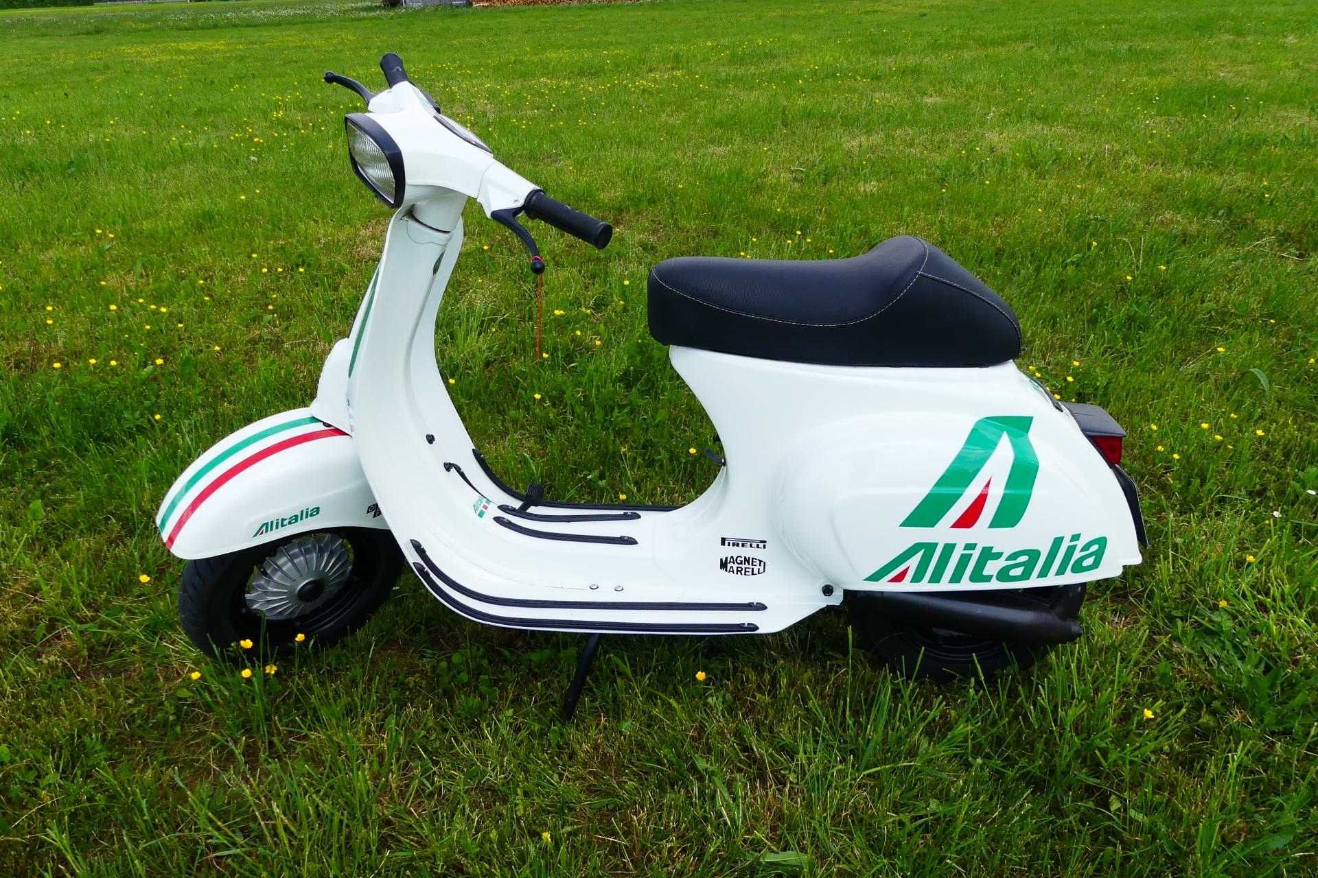 Ein weißer Motorroller mit Alitalia-Schriftzug, geparkt auf saftig grünem Gras mit kleinen gelben Blumen.