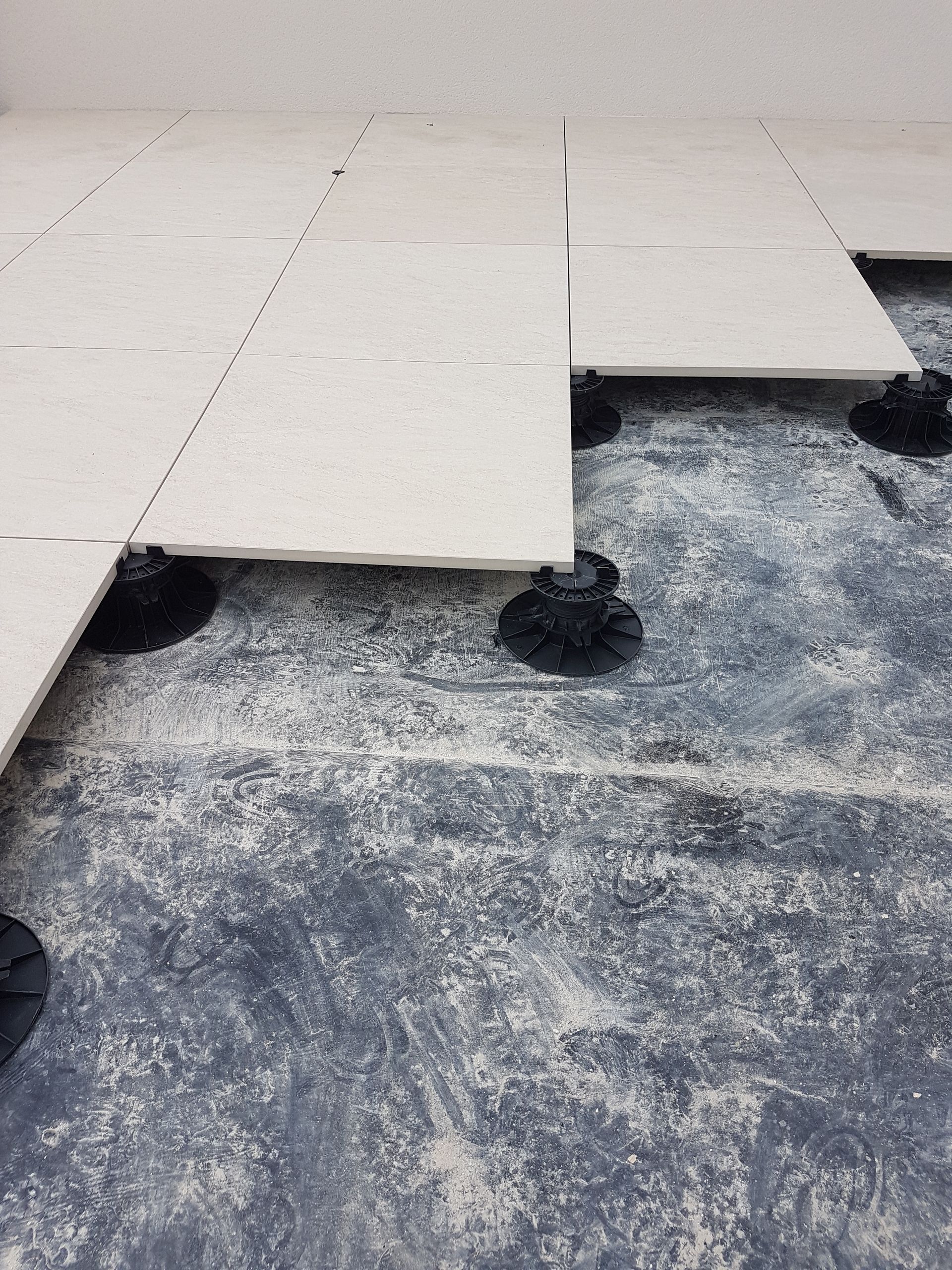 Des carreaux de sol blancs non fixés, avec des croisillons noirs, posés sur un sous-plancher foncé, montrent une installation de revêtement de sol inachevée.