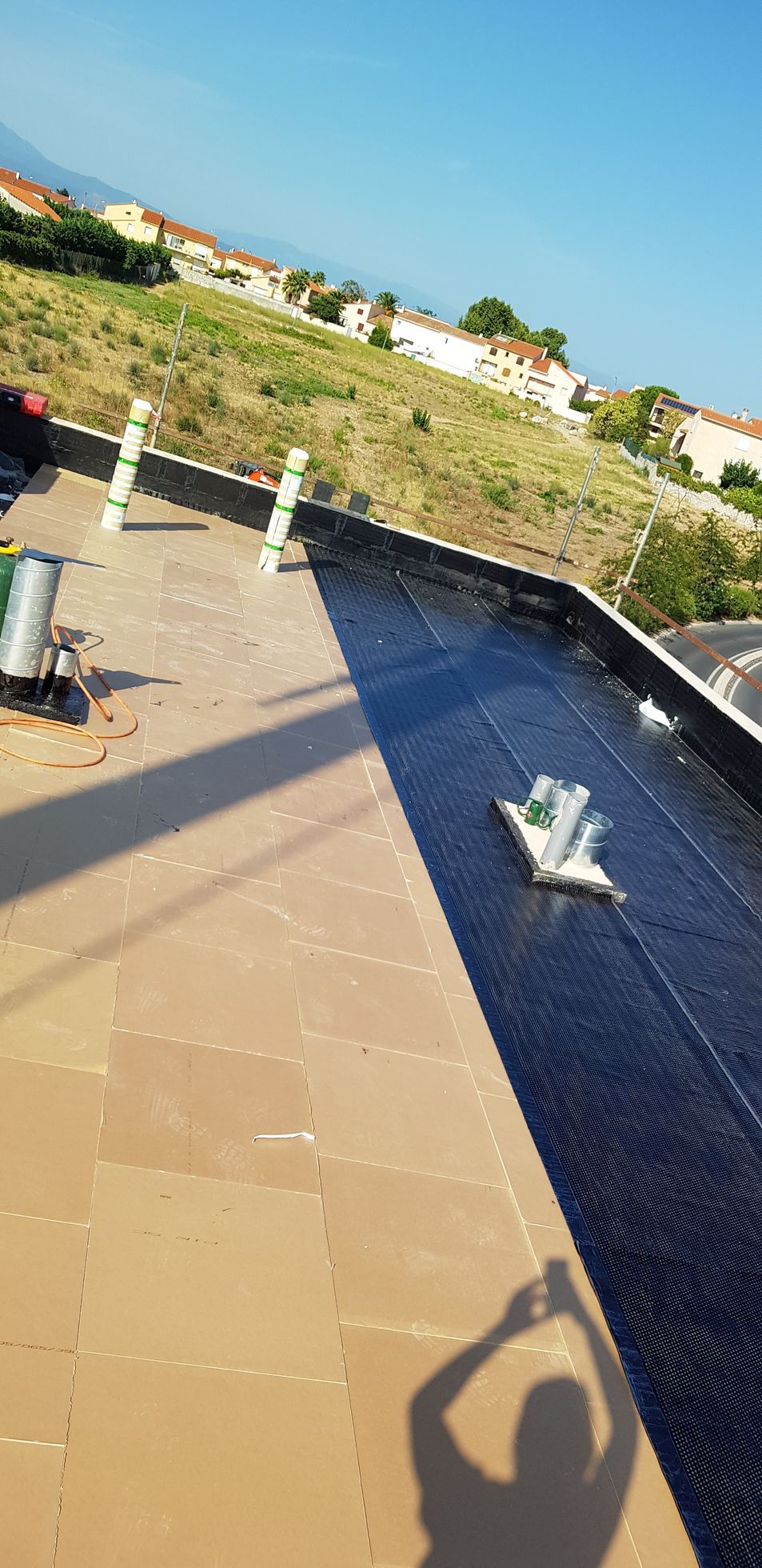 Terrasse sur le toit avec puits de lumière et une personne dans l'ombre prenant une photo, donnant sur une colline herbeuse et des maisons