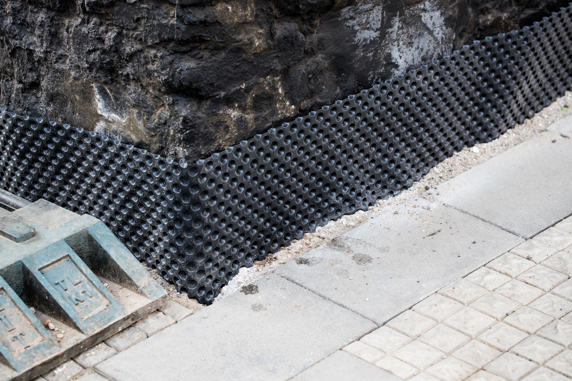 Tapis de drainage noir sur une pente rocheuse, à côté d'une allée pavée gris clair et d'une grille d'égout.