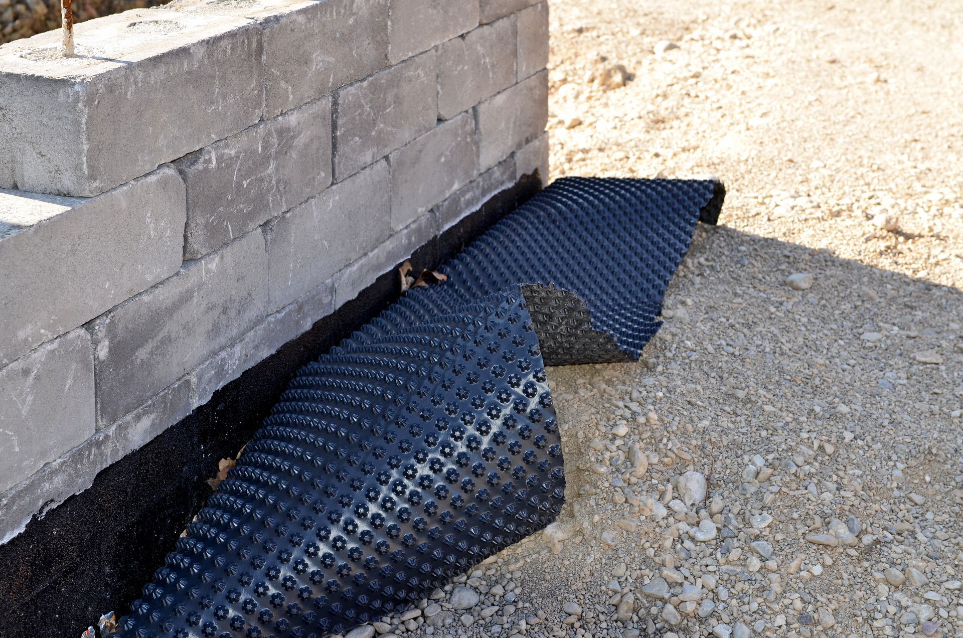 Tapis de drainage noir partiellement dissimulé sous un mur en parpaings sur un sol en gravier