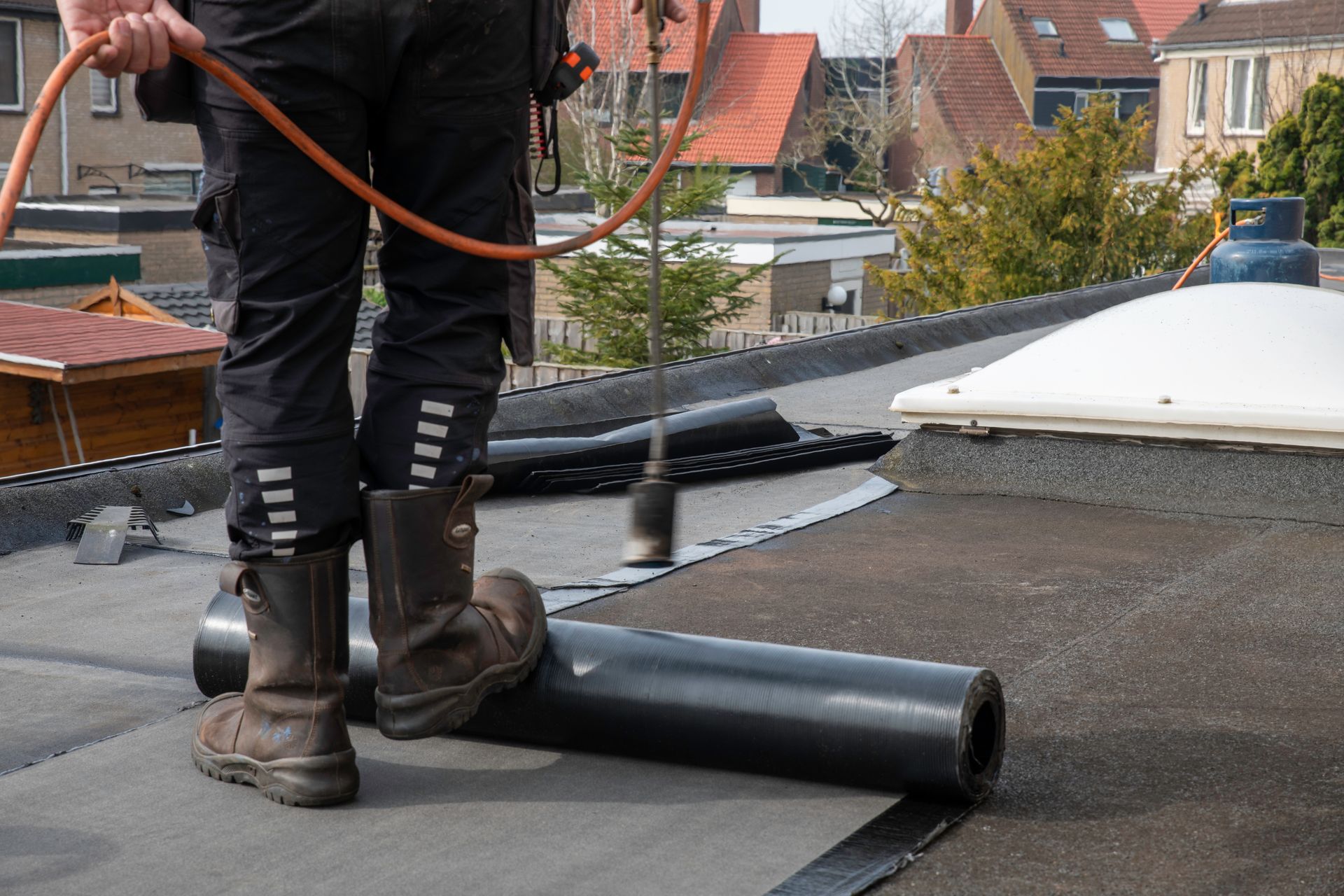 Ouvrier installant une membrane de toiture noire sur un toit plat, le tuyau d'un chalumeau thermique étant visible.