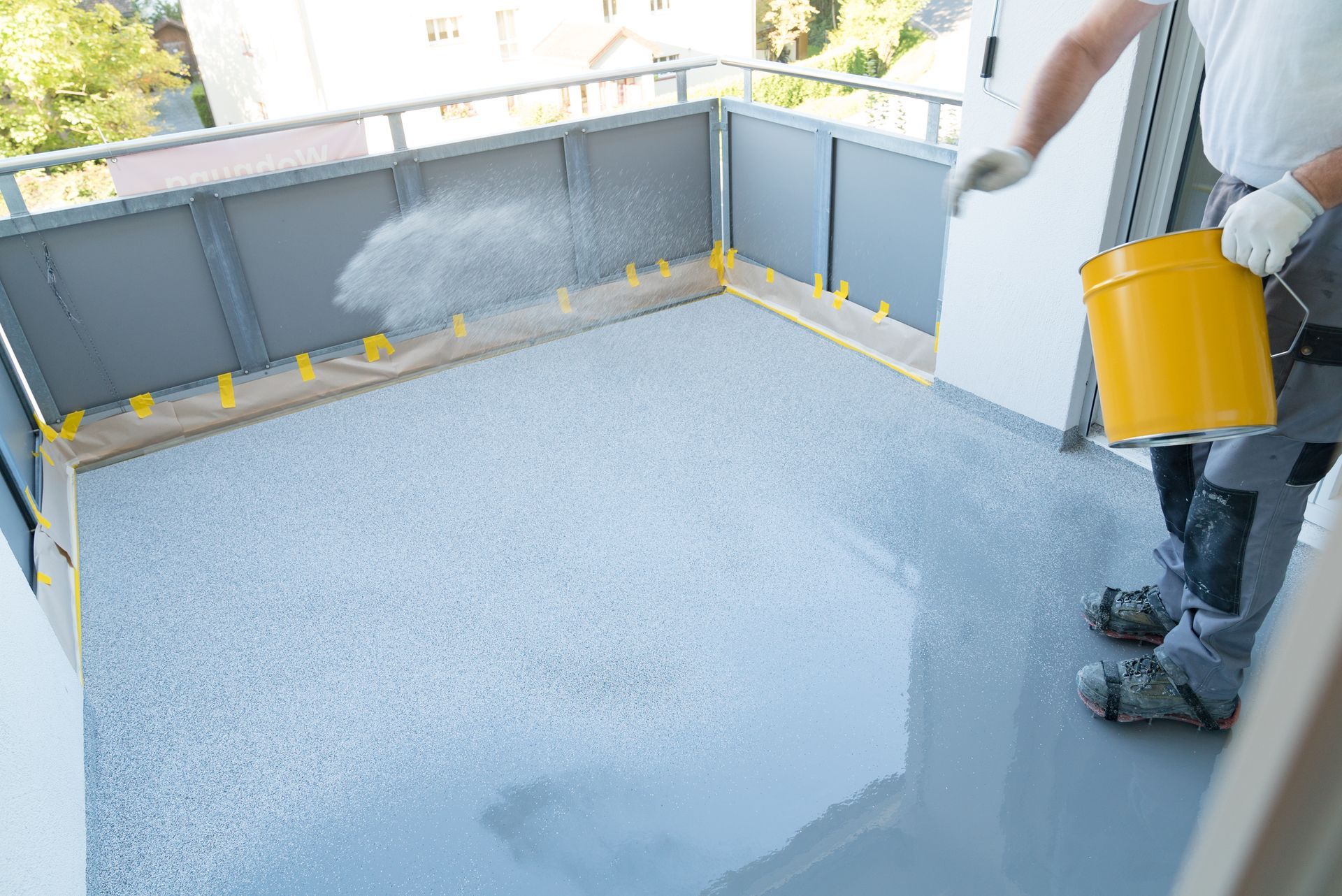 Une personne applique un revêtement gris brillant sur le sol d'un balcon à l'aide d'un seau jaune.