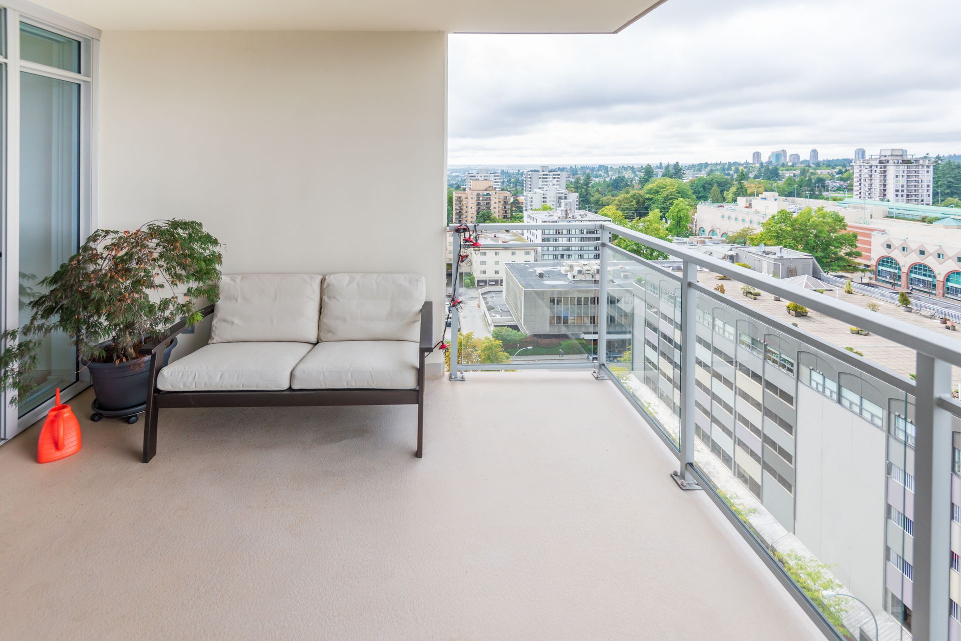 Balcon avec un canapé deux places gris, une plante en pot et une vue sur la ville au-delà d'une balustrade en verre.