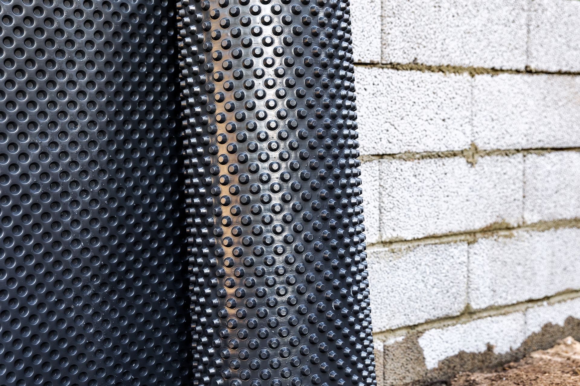 Membrane de drainage noire à picots contre un mur en parpaings blancs avec un soubassement en terre.