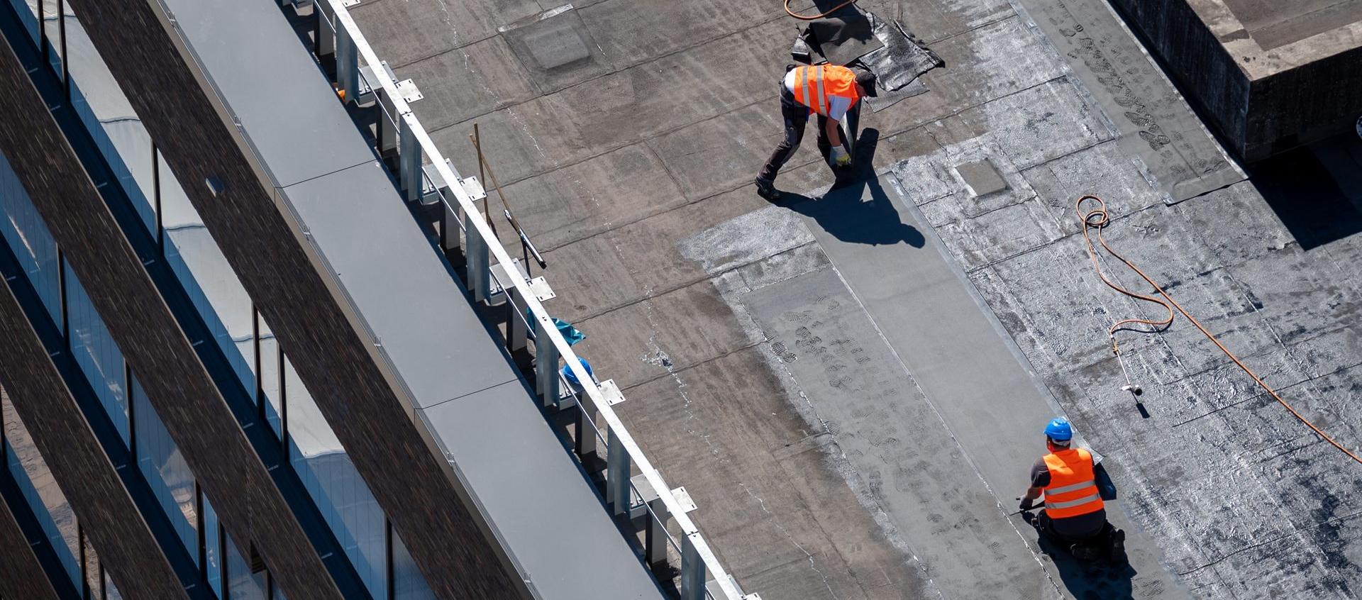 Des ouvriers du bâtiment en gilets orange travaillent sur un toit, près du bord d'un bâtiment.