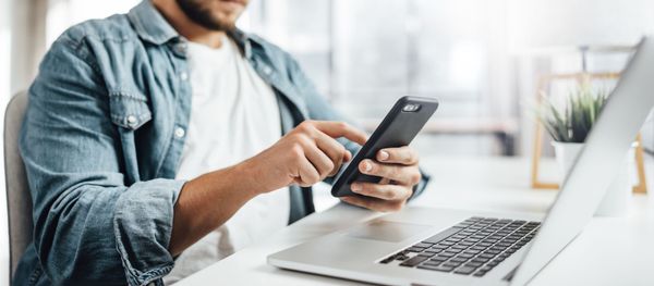 Une personne utilise un smartphone à côté d'un ordinateur portable ouvert, à un bureau dans un bureau lumineux.