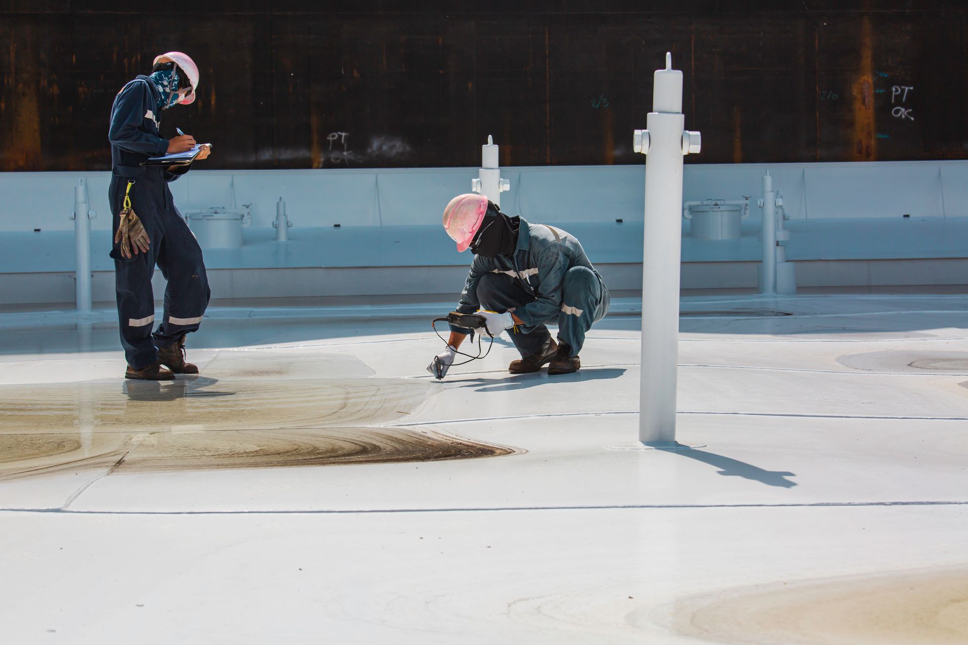 Deux ouvriers portant des casques de chantier inspectent un toit mouillé avec des tuyaux d'aération blancs et un tuyau d'arrosage qui projette de l'eau.