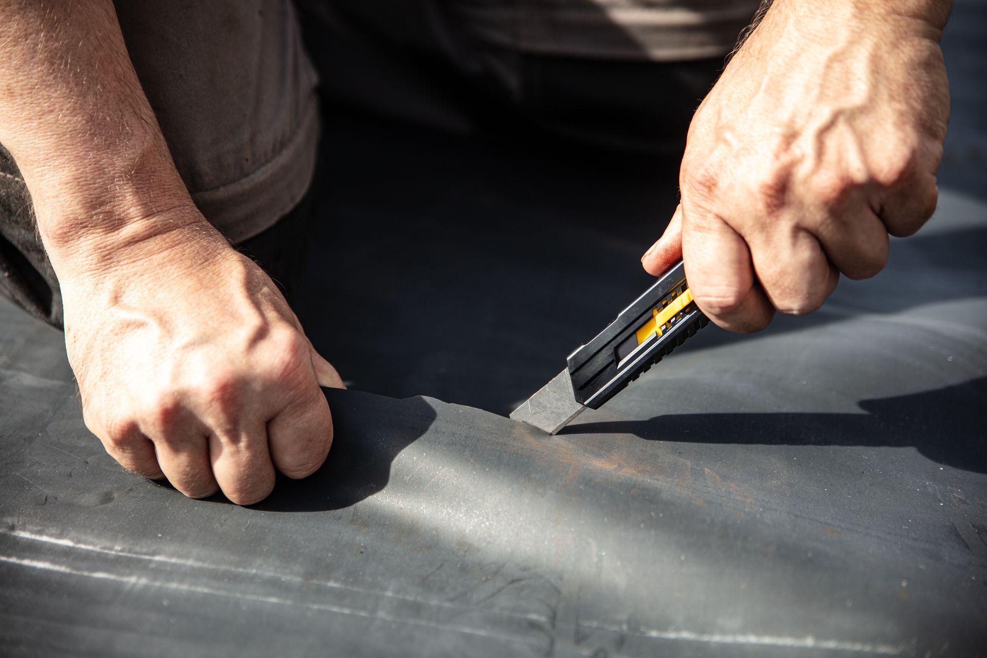 Des mains utilisent un couteau utilitaire pour couper un matériau de toiture noir sur une surface plane