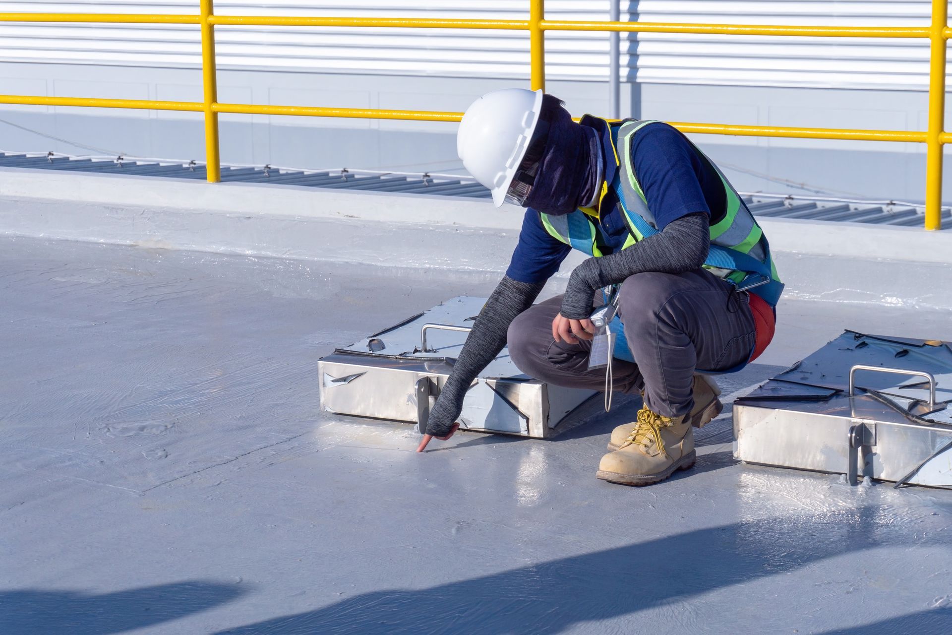 Un ouvrier portant un casque de chantier inspecte du matériel sur un toit muni d'une rambarde de sécurité jaune.