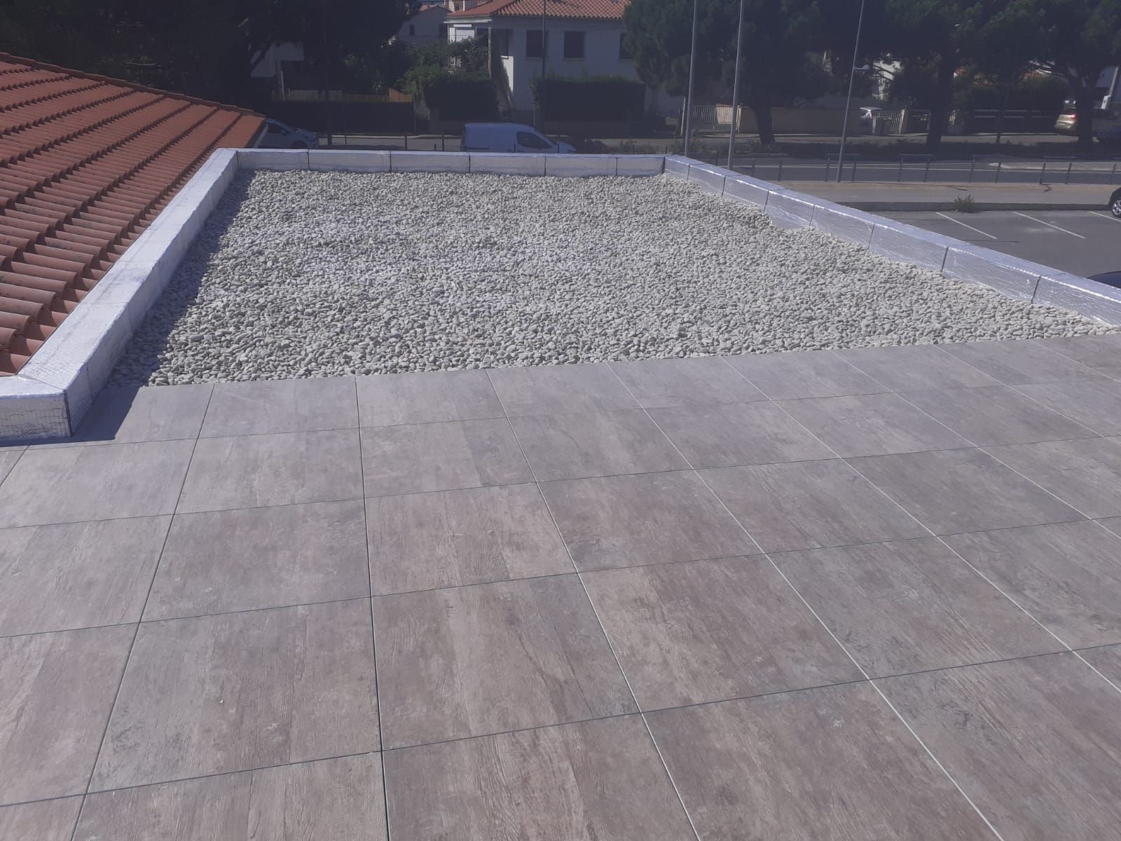 Toiture-terrasse avec dalles grises et une partie en gravier, jouxtant des toits de tuiles rouges et des maisons au loin.