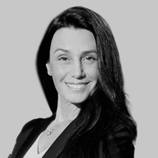 Mujer sonriente con cabello largo y oscuro, vestida con un blazer negro, sobre un fondo gris claro.