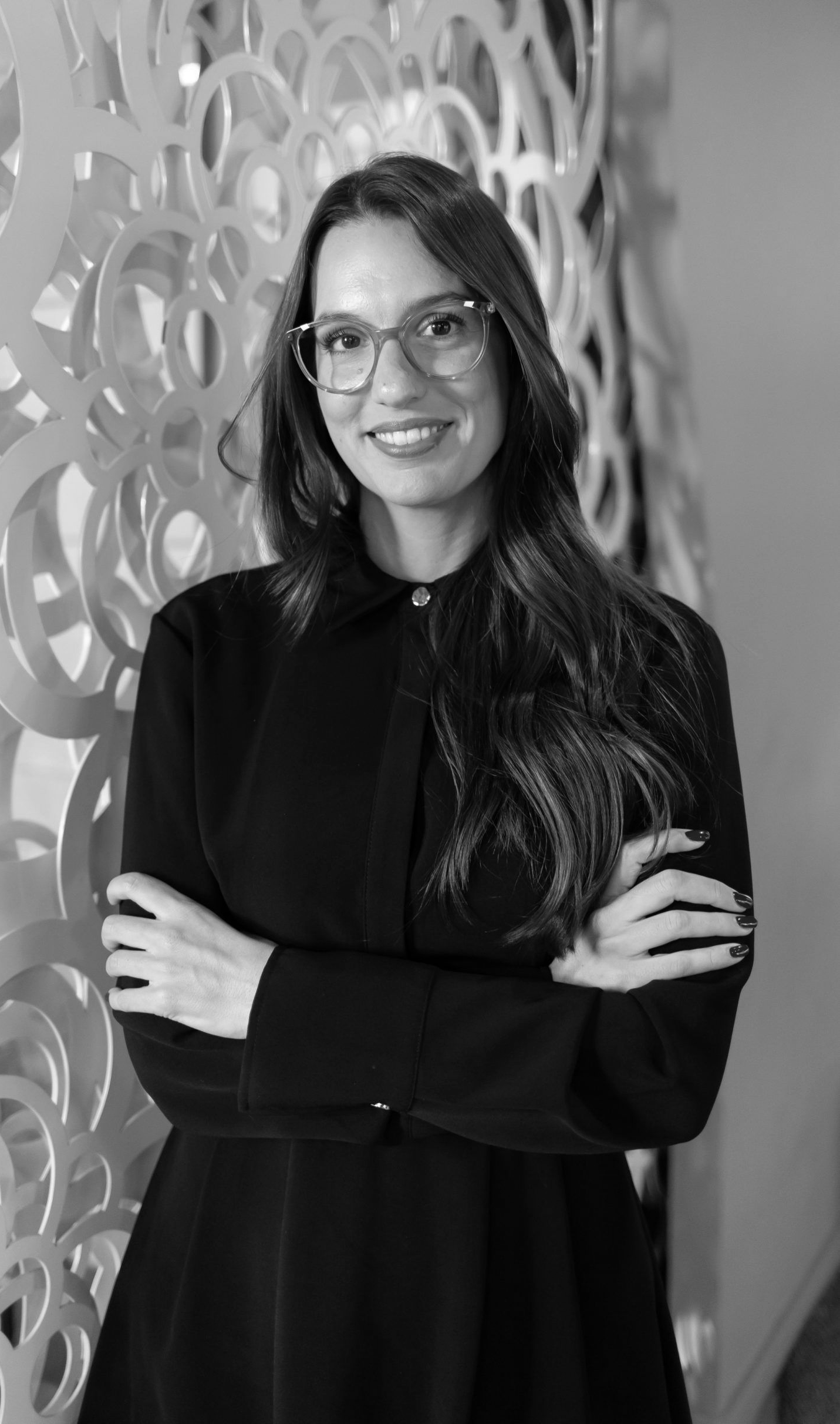 Una persona con cabello largo castaño y gafas sonríe con los brazos cruzados, vistiendo una camisa negra