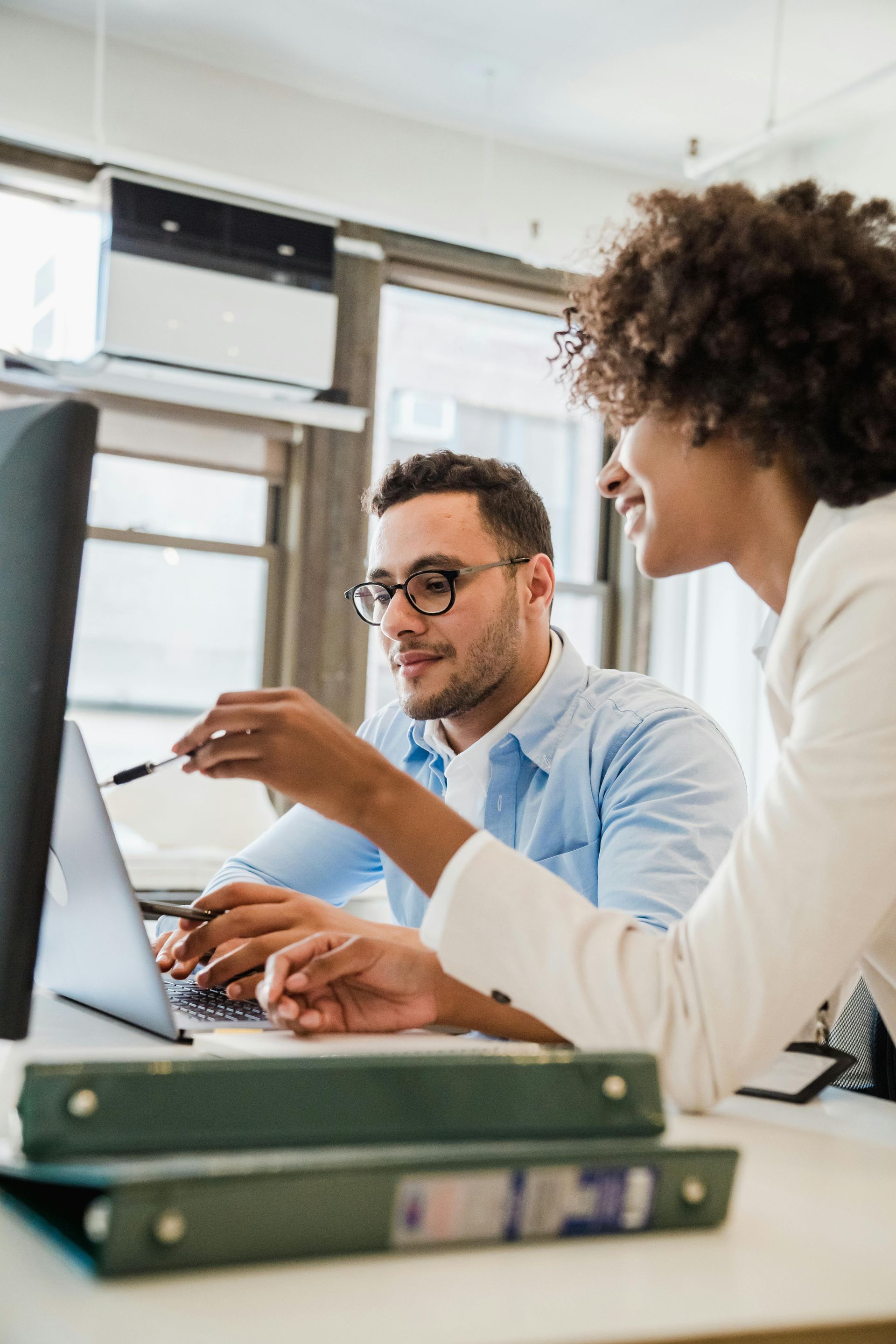 Dos compañeros de oficina colaboran frente a un ordenador; uno señala la pantalla mientras el otro observa.