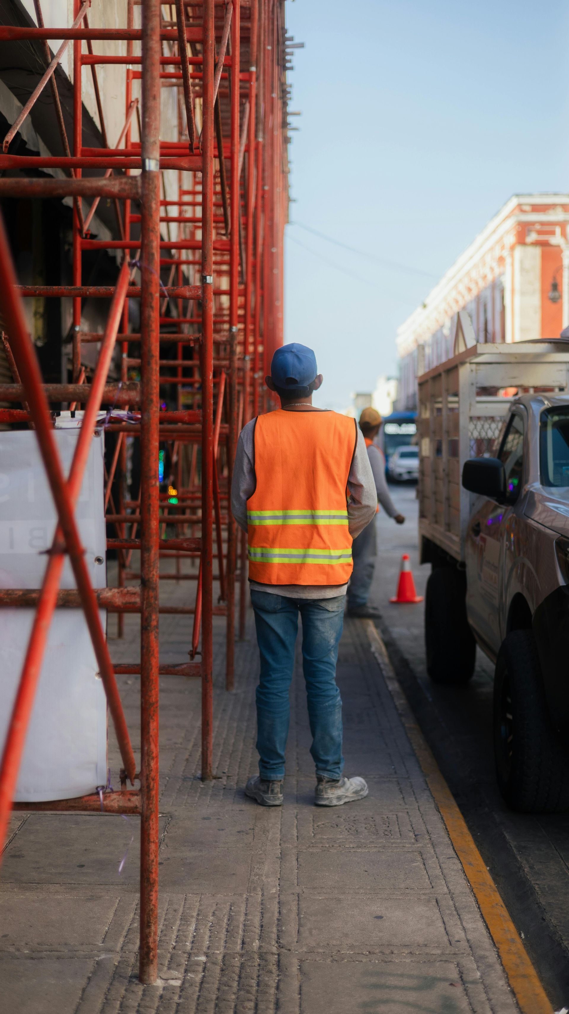 Una persona con un chaleco naranja de alta visibilidad y un casco azul se encuentra de pie en una acera de la ciudad