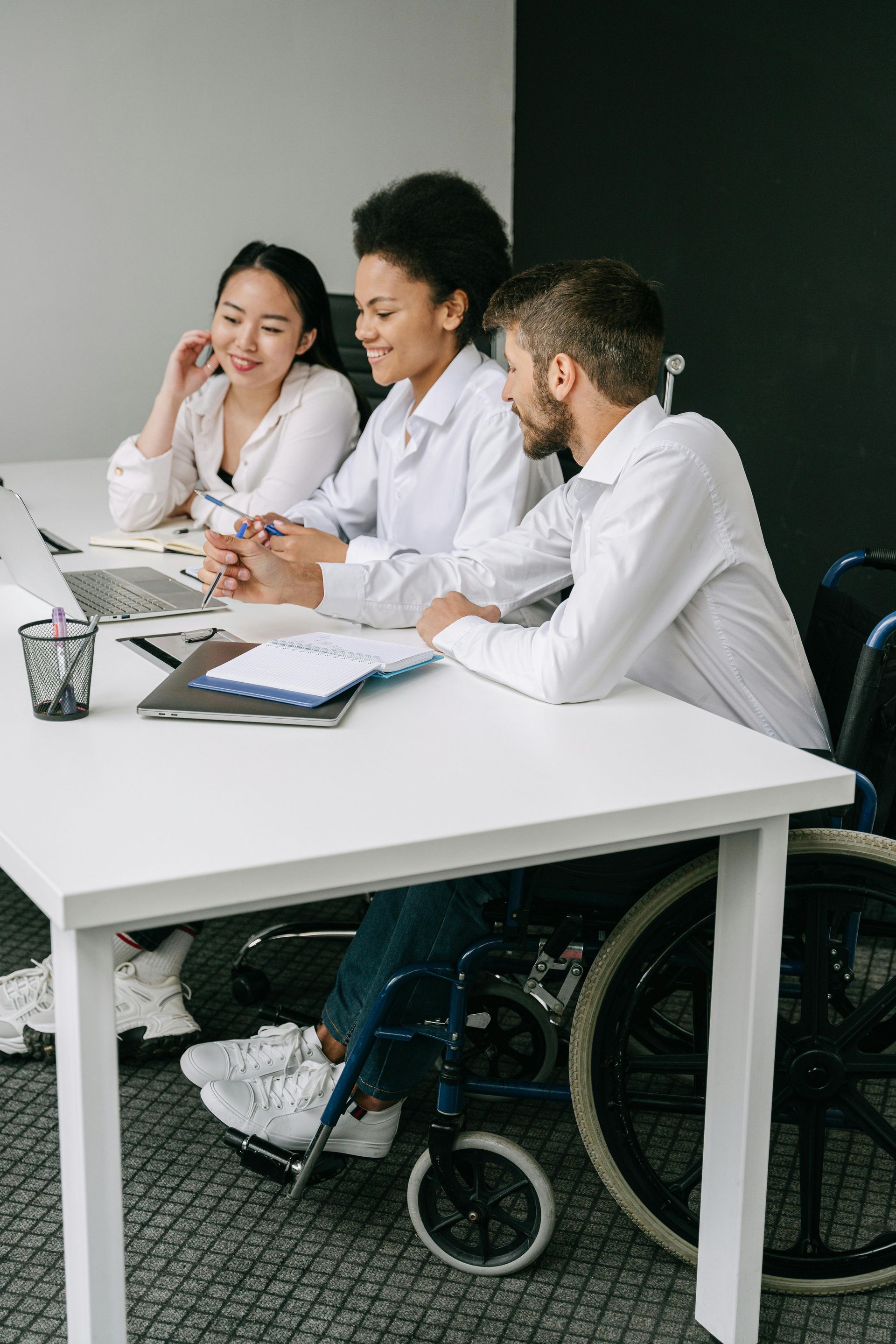 Tres personas están sentadas en una mesa blanca en una oficina, hablando de trabajo mientras miran una computadora portátil