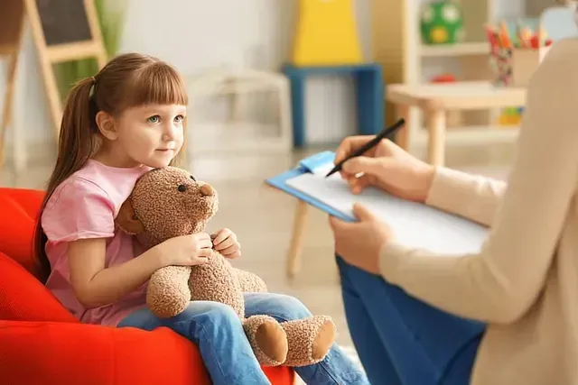 Una niña está sentada en una silla roja sosteniendo un osito de peluche y hablando con un psicólogo infantil.