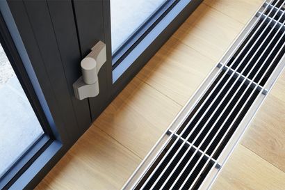 Türgriff und Bodenlüftungsdetails neben einem Fenster auf hellem Holzboden. Die Tür ist dunkelgrau.