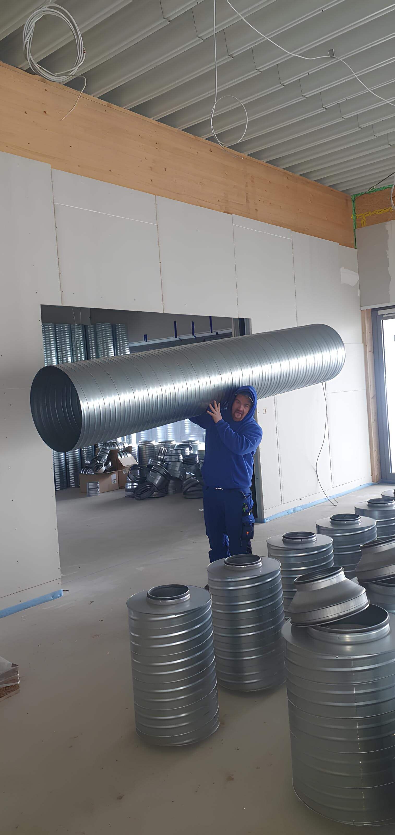 Ein Bauarbeiter trägt ein großes Metallrohr. Im Inneren des Gebäudes liegen weitere Rohre auf dem Boden.