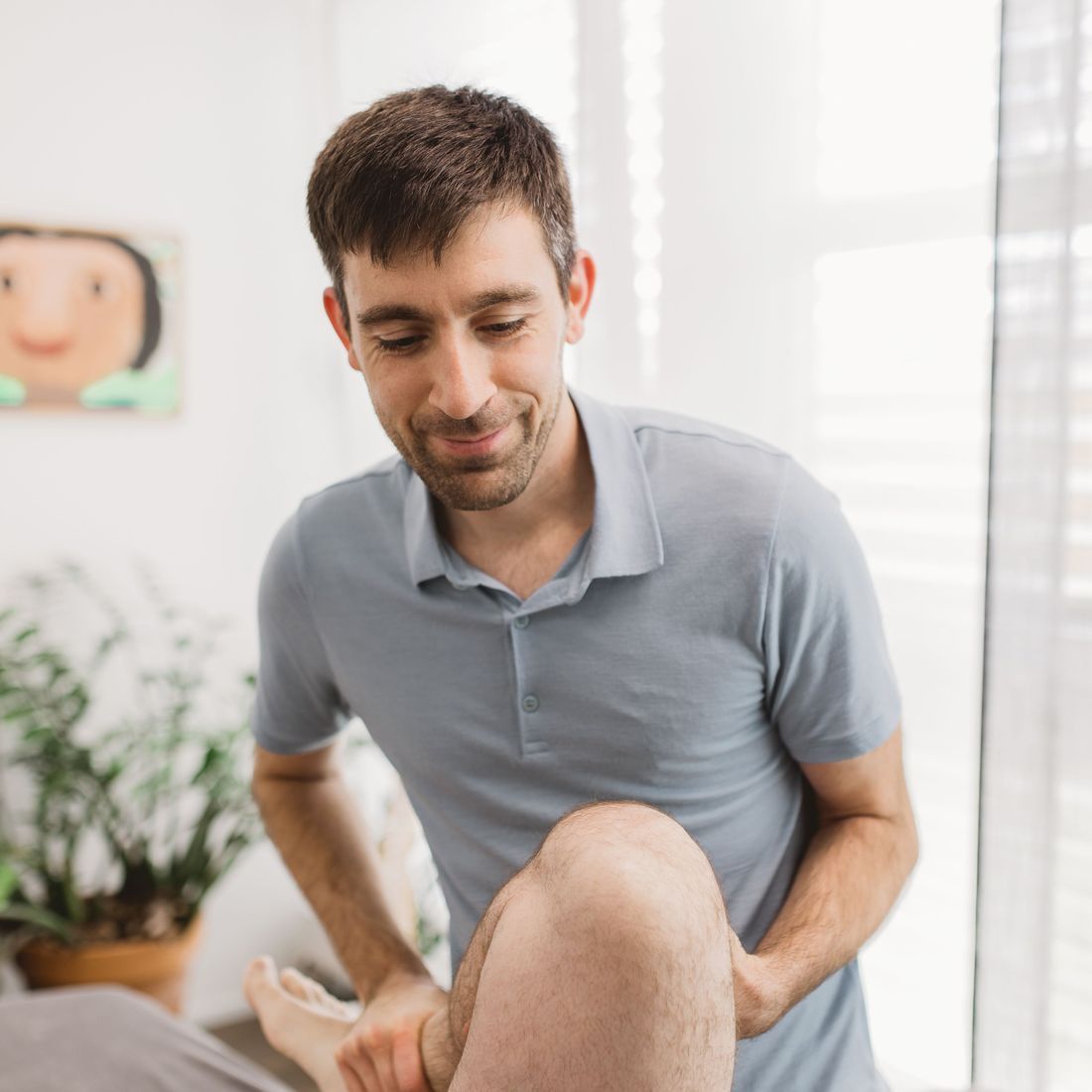 Ein männlicher Therapeut in einem grauen Hemd untersucht in einem hell erleuchteten Raum das Bein eines Patienten.