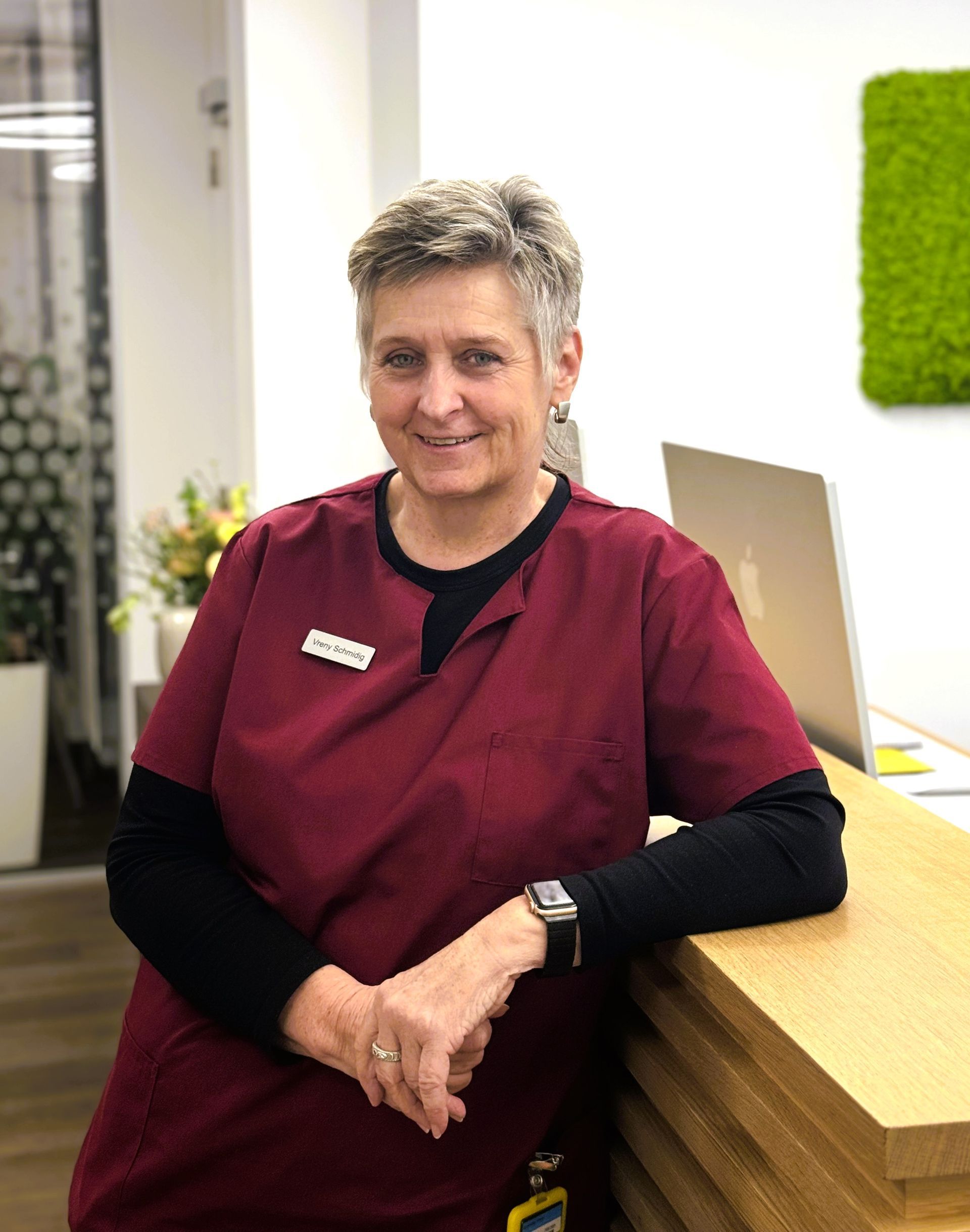 Eine Frau in OP-Kleidung lehnt an einem Holztresen. , Sie lächelt. Hinter ihr befindet sich ein modernes Büro.