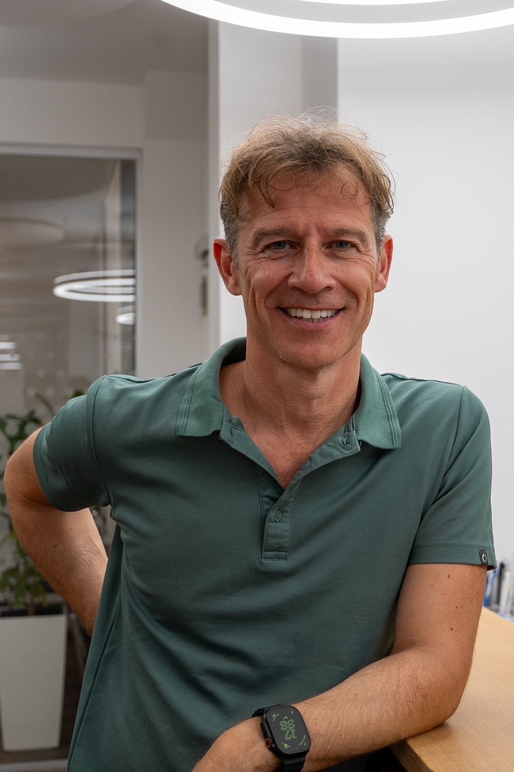 Ein Mann mit Brille und rotem Hemd sitzt mit einem Stethoskop an einem Schreibtisch und lächelt in die Kamera.