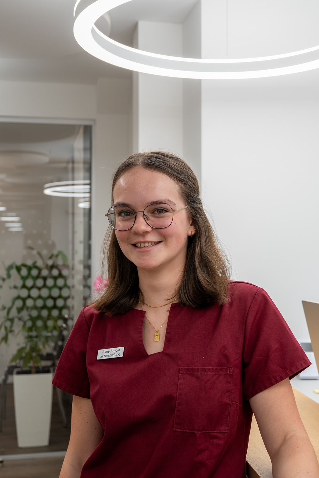 Lächelnde Frau in OP-Kleidung an einem Empfangstresen mit einem Laptop und einem grünen Moosquadrat an der Wand.