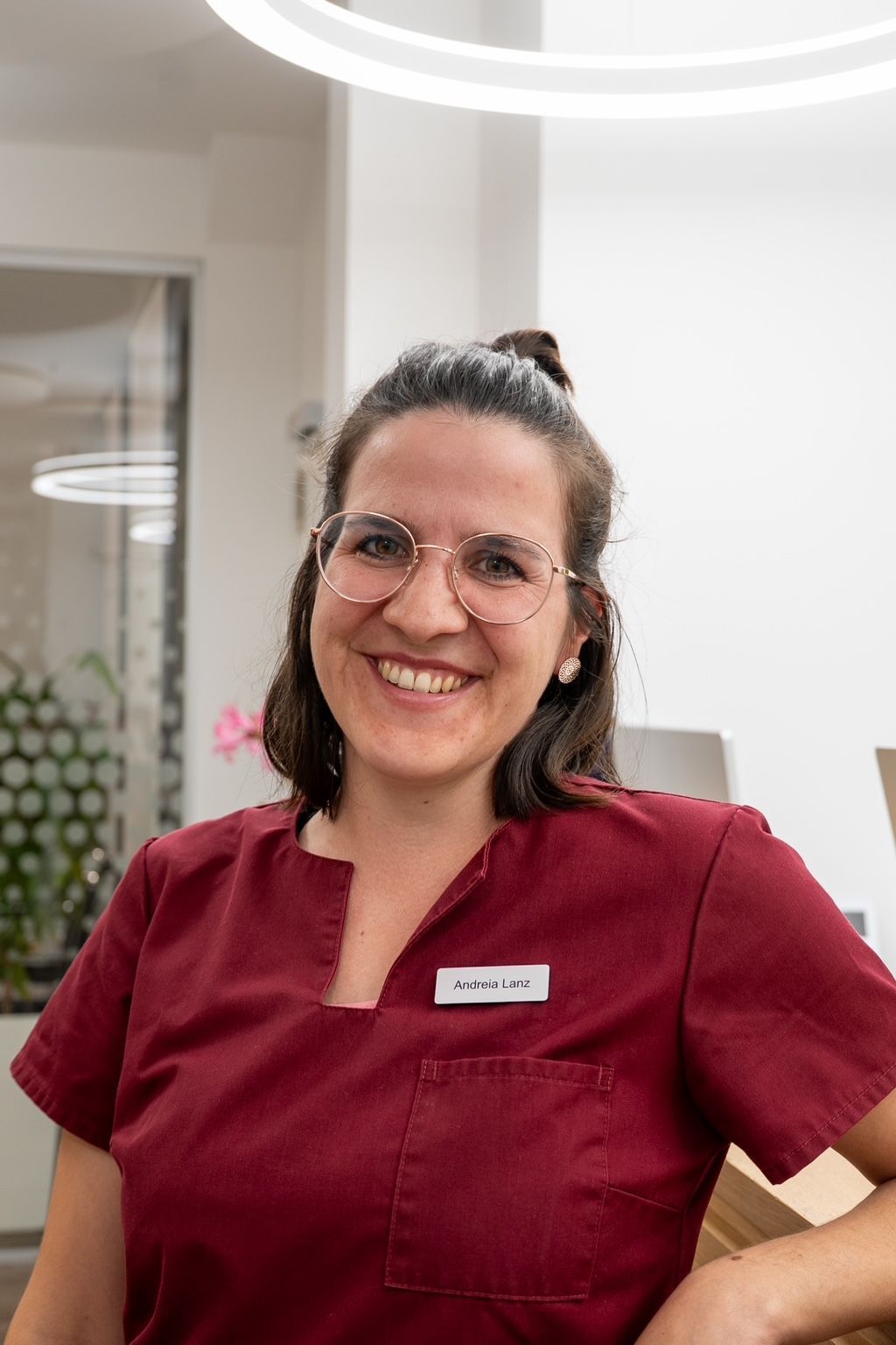 Dame in OP-Kleidung lehnt lächelnd an einem Schreibtisch in einem hellen Büro. Sie trägt eine Brille und ein Namensschild.