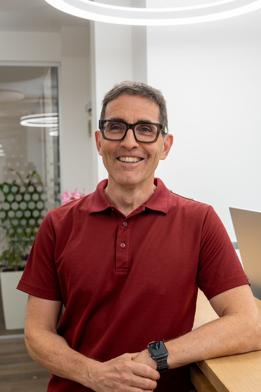 Mann mit Brille und rotem Poloshirt lehnt lächelnd an einer Holztheke. Büroumgebung mit Laptop und Mooswand.