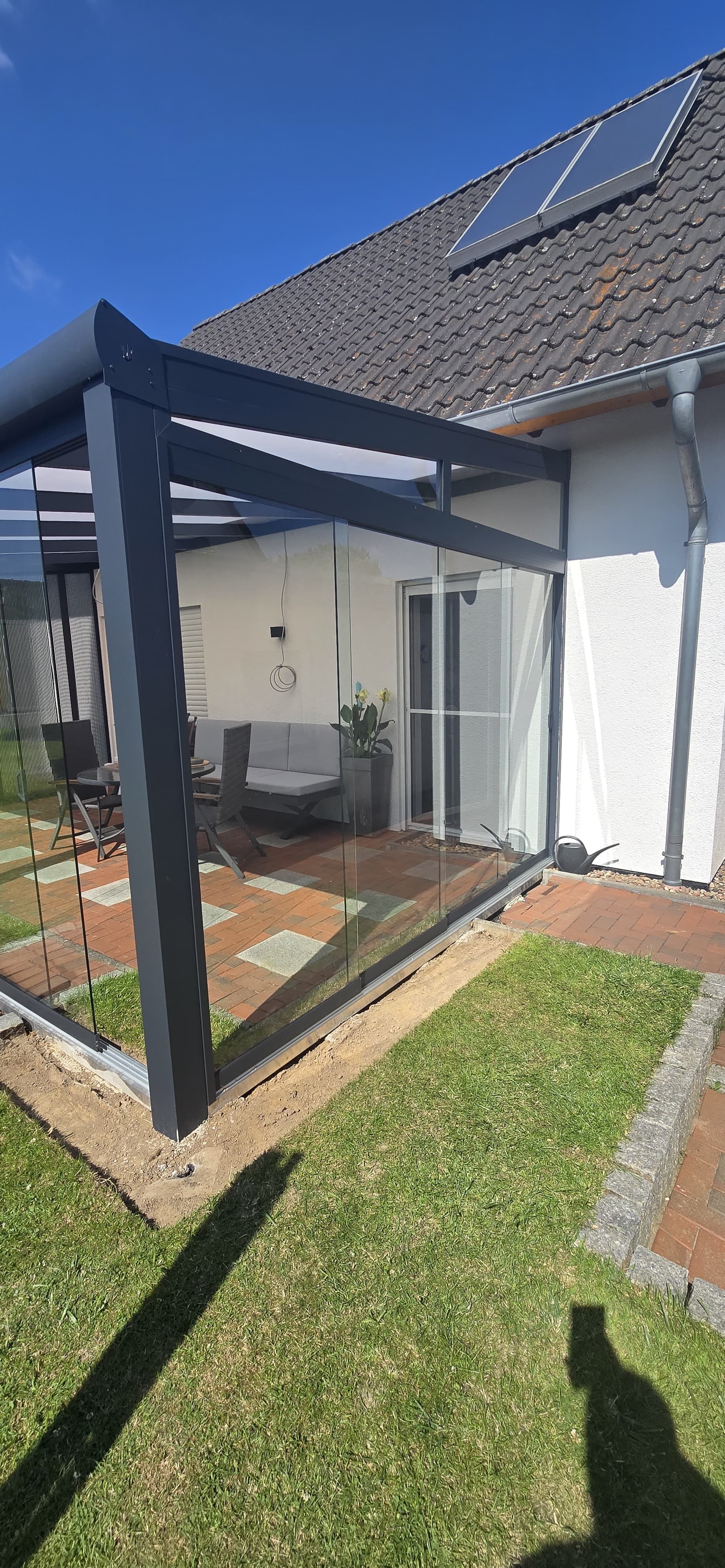 Eine moderne Terrasse mit dunkelgrauem Rahmen, klaren Glaswänden und Glasdach. Die Terrasse hat einen Steinboden und ist von grünem Gras umgeben.