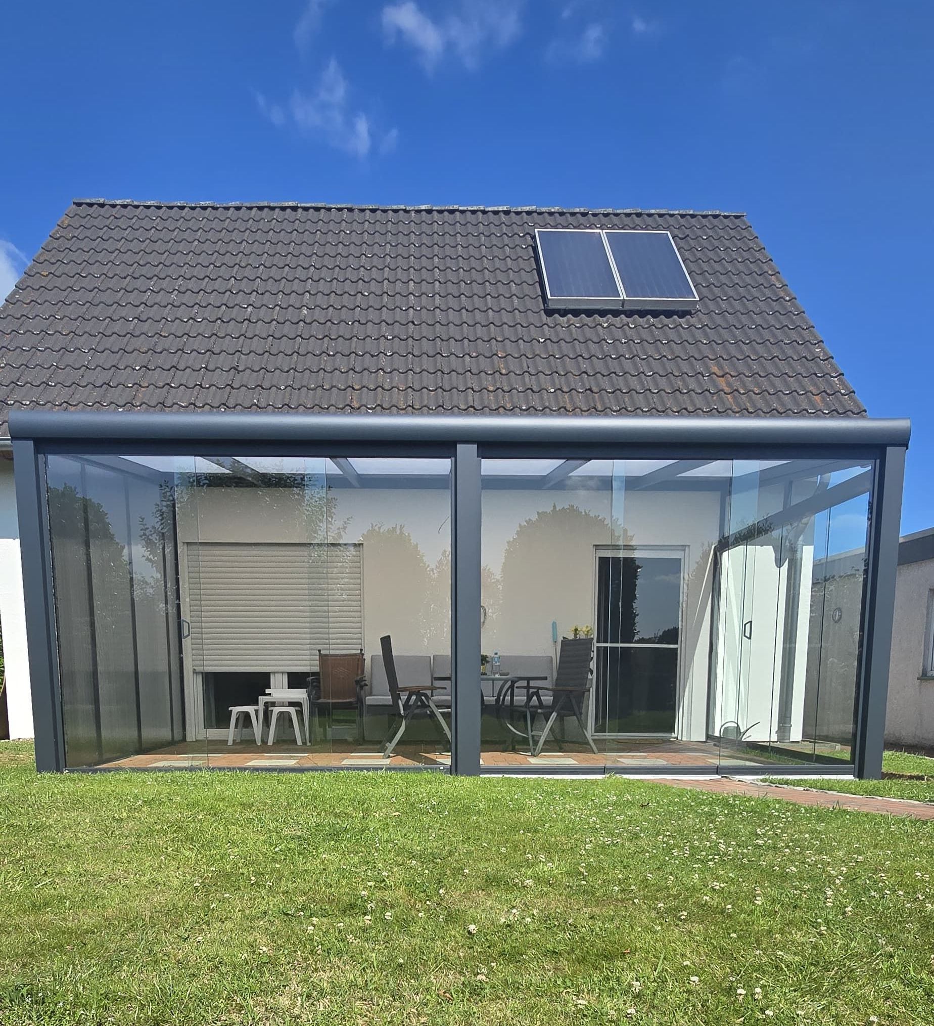 Verglaste Terrasse an einem Haus. Dunkelgraues Dach mit Solarpanel, Rasen, durch das Glas sichtbare Essmöbel im Freien.