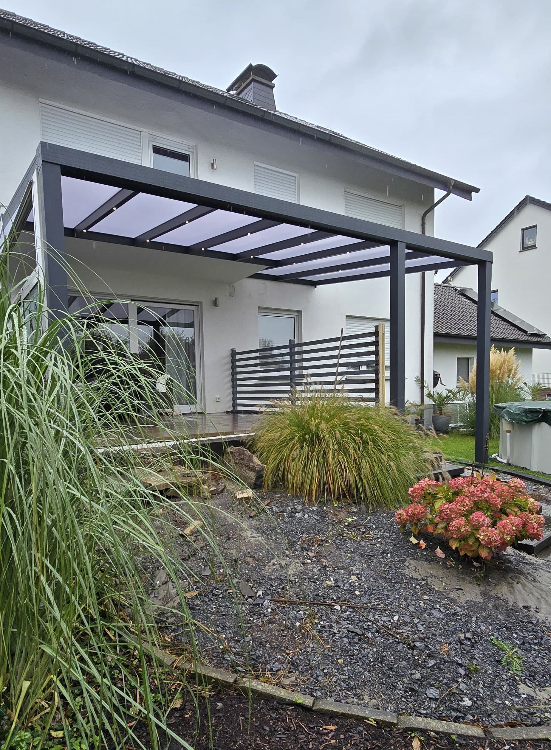 Schwarze Pergola, die eine Terrasse bedeckt, die an ein weißes Haus angrenzt; dunkelgraue Kieslandschaft.