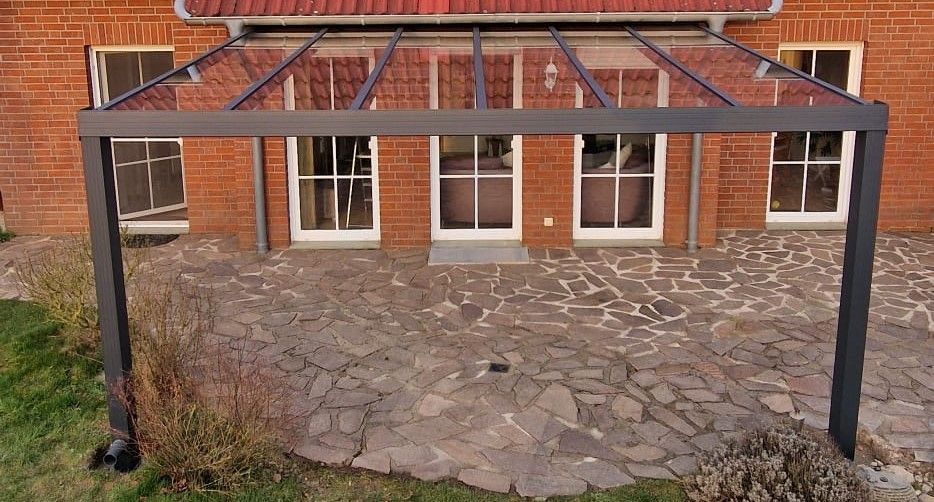 Graue Pergola über einer Steinterrasse vor einem Backsteinhaus mit Glastüren und -fenstern.