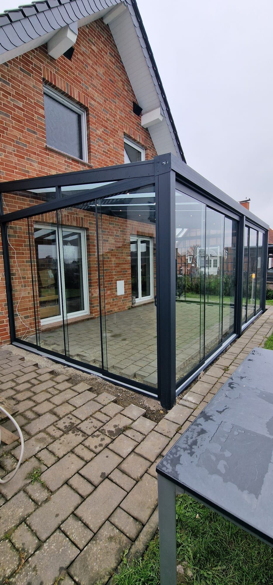 Backsteinhaus mit verglaster Terrasse. Schwarzer Rahmen, klares Glas, Backsteinterrasse und bewölkter Himmel.