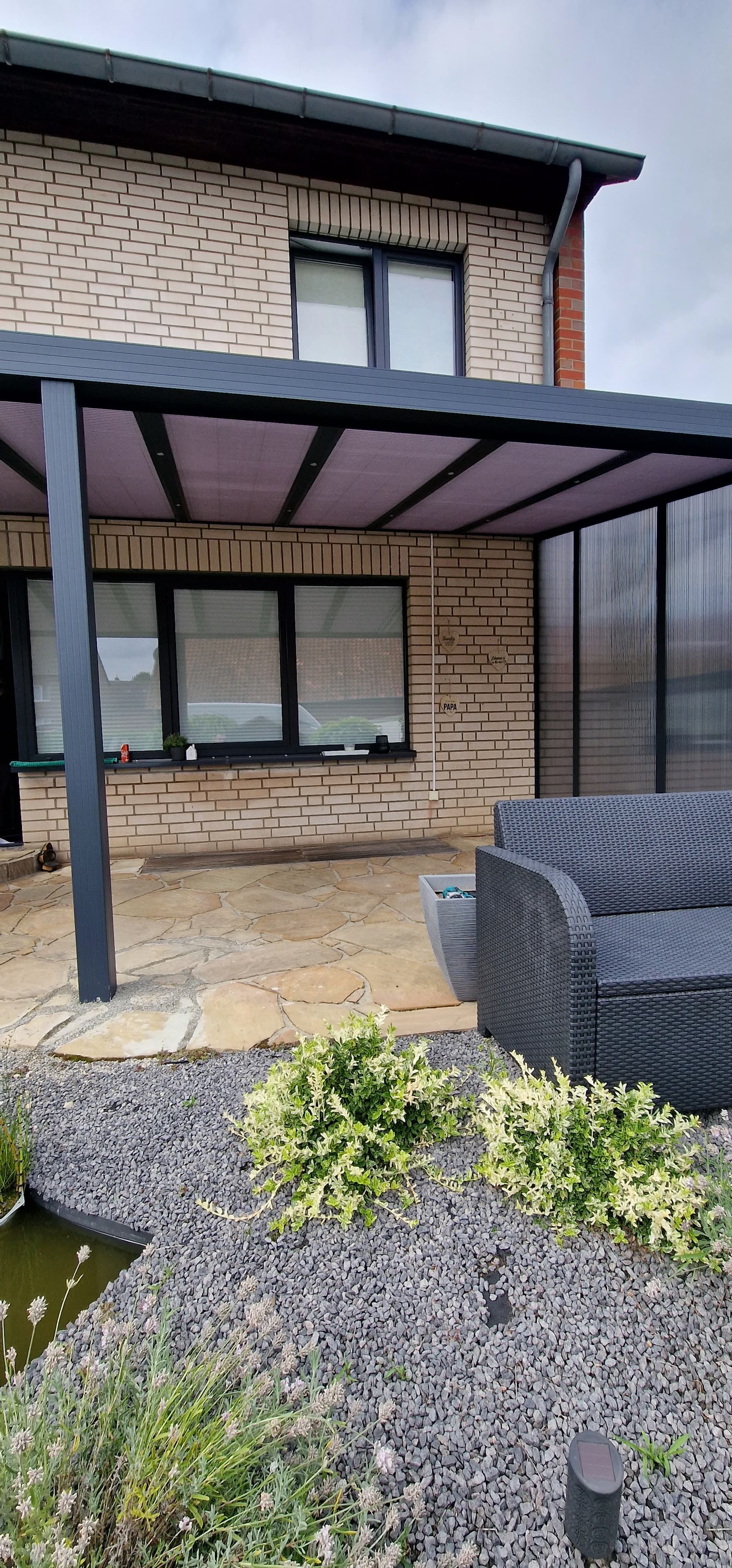 Pergola über einer gemauerten Terrasse. Ein kleiner Steingarten mit Teich im Vordergrund.