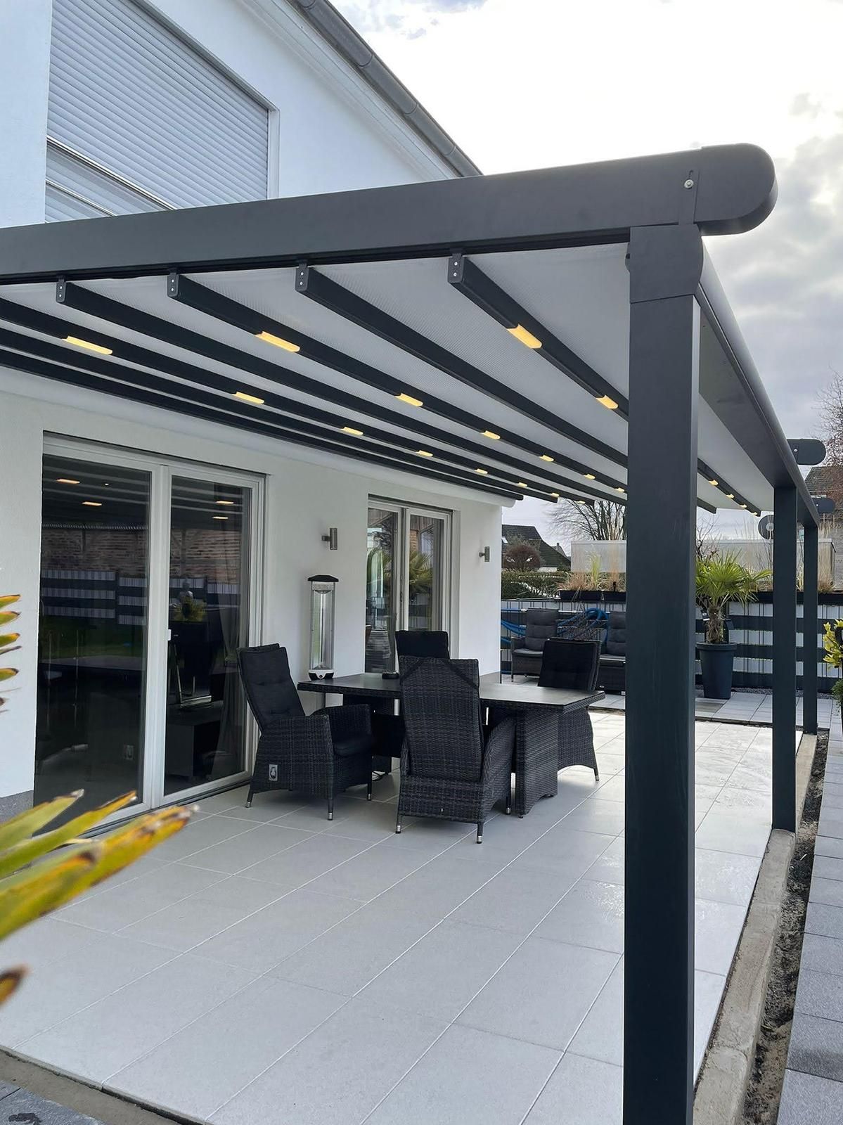 Essbereich im Freien mit dunkelgrauer Pergola mit Beleuchtung auf weißer Terrasse mit Möbeln.