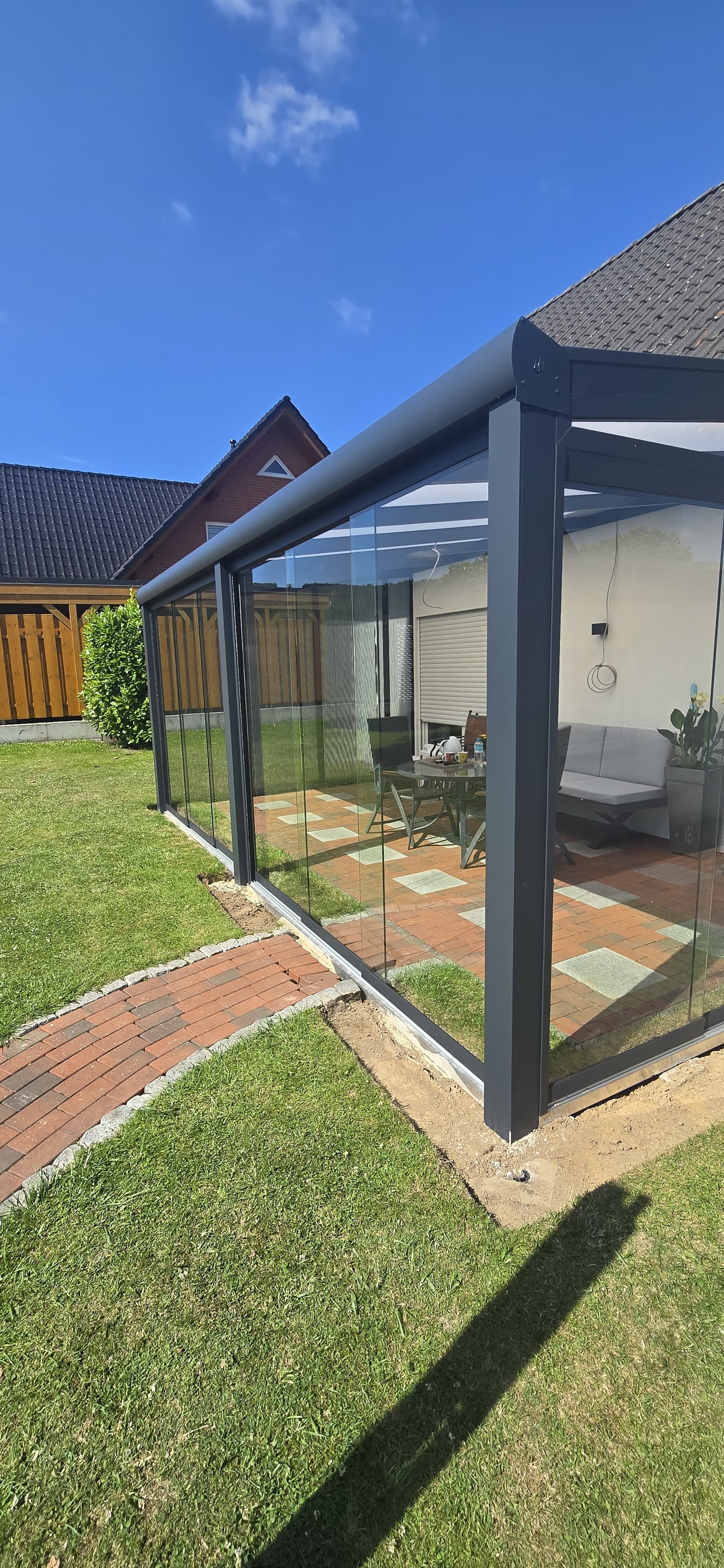Eine moderne graue Terrassenüberdachung mit Glasplatten in einem Hinterhof, einem Ziegelweg und einem grünen Rasen. Hintergrund: blauer Himmel.