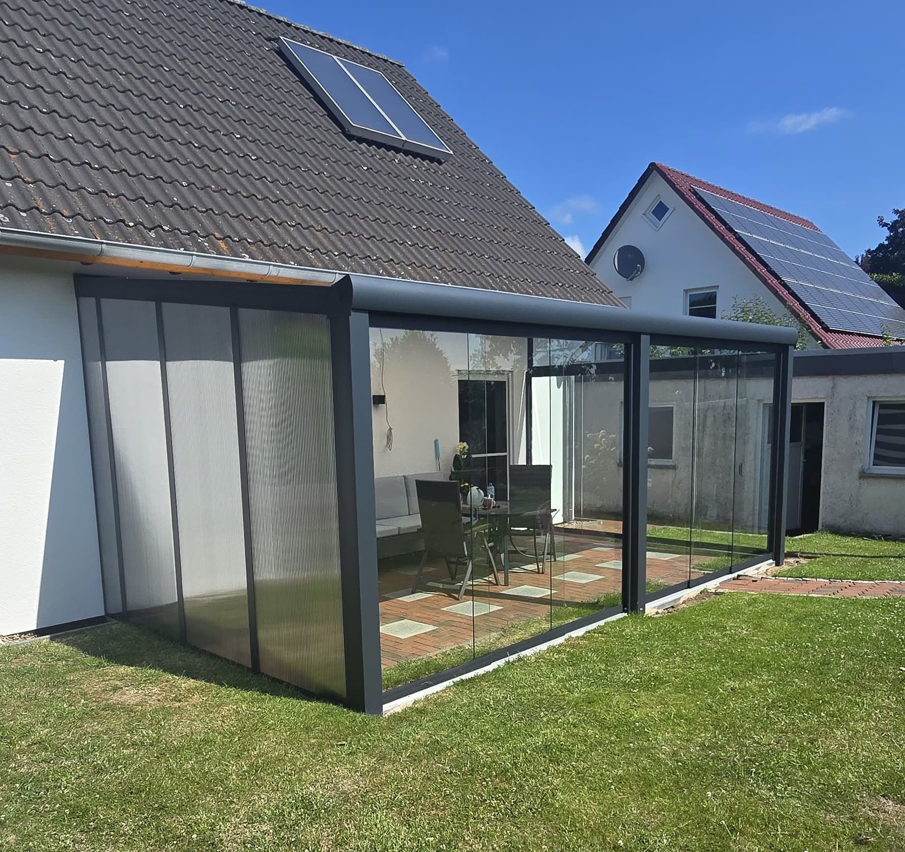 Geschlossene Terrasse an einem Haus mit Glaswänden, grauem Rahmen und einem Sitzbereich im Freien auf einer gemauerten Terrasse.