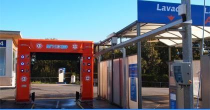 Entrada de un autolavado de autoservicio rojo, con letreros azules y una zona de lavado al aire libre con techo