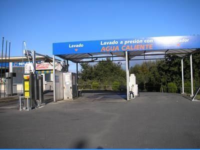 Entrada de un lavadero de coches con marquesina azul y carteles en español sobre el lavado a presión con agua caliente